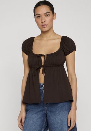 Kvinde iført en sort kortærmet top med bindebånd foran og blå denimjeans, stående mod en ensartet lys baggrund.