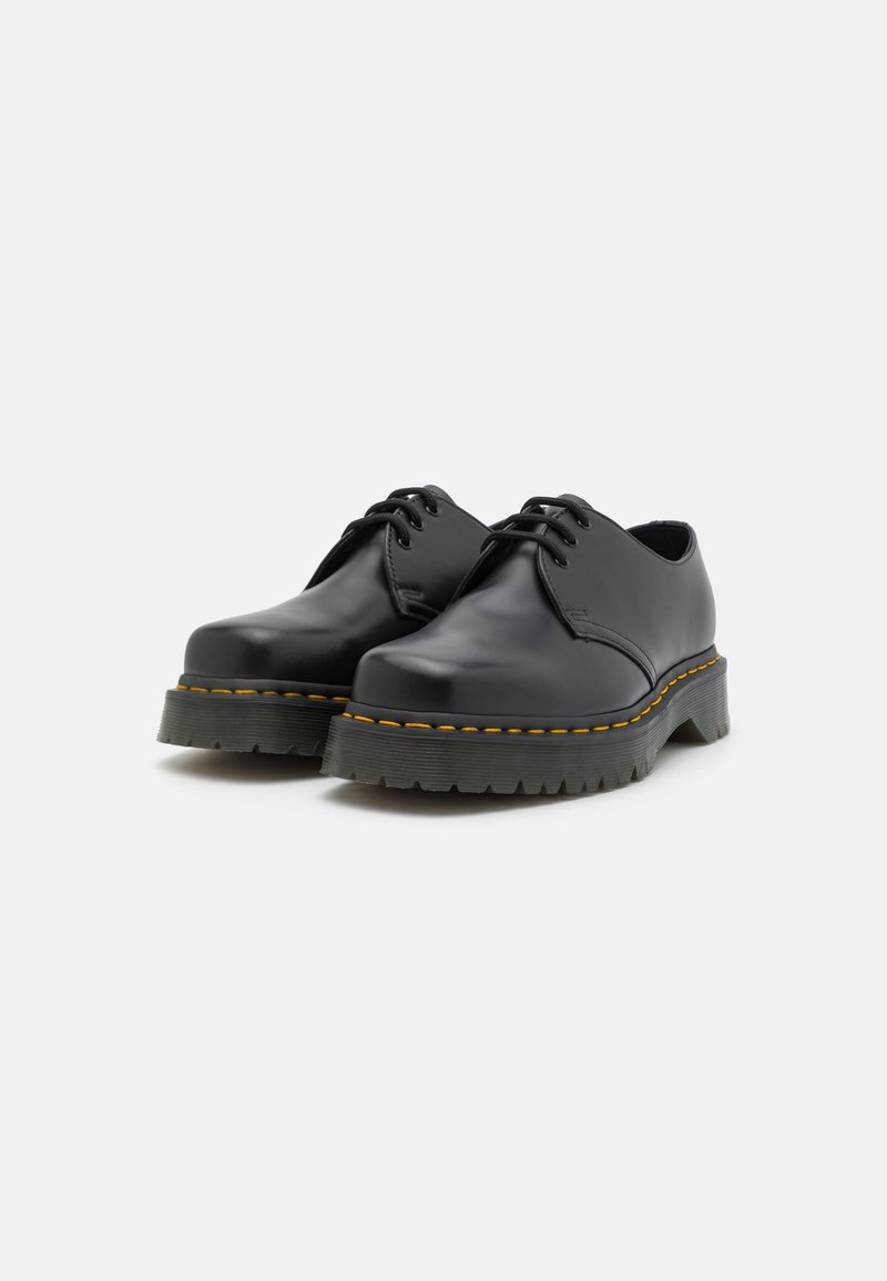 Zapatos de cuero negro con cordones, punta redondeada, suela gruesa de goma negra y costuras amarillas distintivas a lo largo del borde.