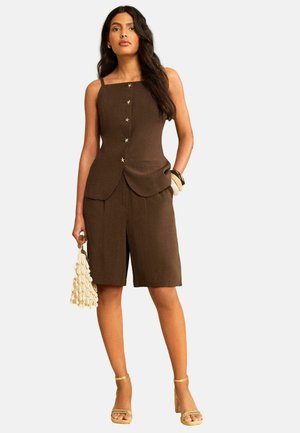 Femme portant un haut marron sans manches boutonné et un short assorti longueur genou, tenant un sac à main crème texturé et portant des talons beiges.