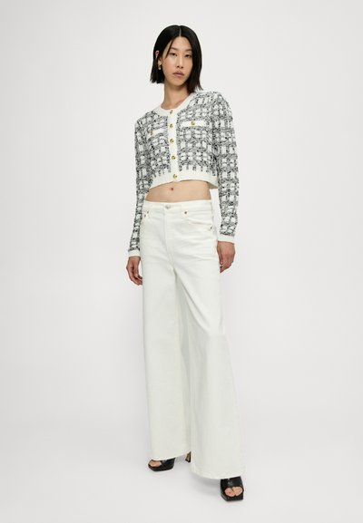 Cardigan à carreaux noir et blanc coupé avec des boutons dorés, associé à un jean évasé blanc et des sandales à talons noires. Textures lisses visibles.