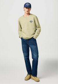 Felpa verde chiaro con il logo "Wrangler Jeans 1947", jeans blu scuro e mocassini beige, completati da un cappellino blu.