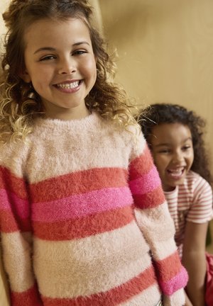 Smilende pige i en blød lyserød og rødstribet trøje, med en anden grinende pige iført en rød- og hvidstribet skjorte i baggrunden.