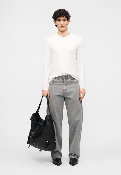 Un tânăr stă cu fața înainte, purtând o cămașă albă cu mâneci lungi, pantaloni gri largi, pantofi negri cu vârf ascuțit și ținând o geantă mare neagră.