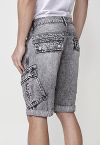 Shorts cargo en denim gris avec un motif décoloré, plusieurs poches latérales, un ourlet retroussé et des détails de couture visibles.