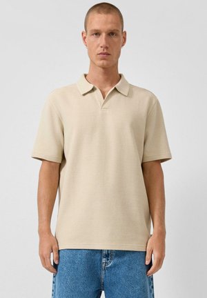 Man in beige poloshirt met korte mouwen en blauwe spijkerbroek, staand tegen een effen lichte achtergrond, met neutrale gezichtsuitdrukking vooruitkijkend.