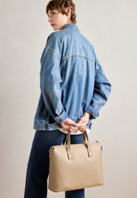 Beige leather tote bag with structured shape, contrasting hardware, and dual top handles, held by a person wearing a denim jacket and jeans.