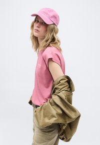 Fiatal hullámos szőke hajú személy rózsaszín sapkát és pólót visel, az egyik karján egy khaki dzsekit tartva, egyszerű fehér háttér előtt.