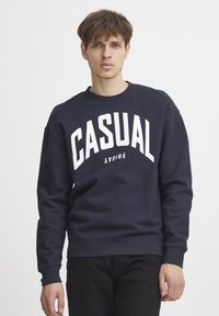Námornícky modrý crewneck sveter vyrobený z bavlneného zmesi, s veľkým bielym textom "CASUAL" na prednej strane, s rebrovanými manžetami a spodným lemom.