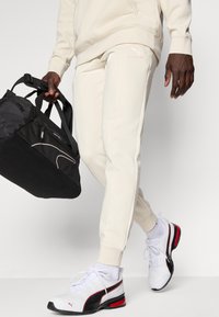 Pantalons de survêtement couleur crème au fit ajusté, avec poches latérales et logo surélevé. Sac de sport noir à bandoulière unique, porté à la main. Baskets blanches avec des accents rouges et noirs.