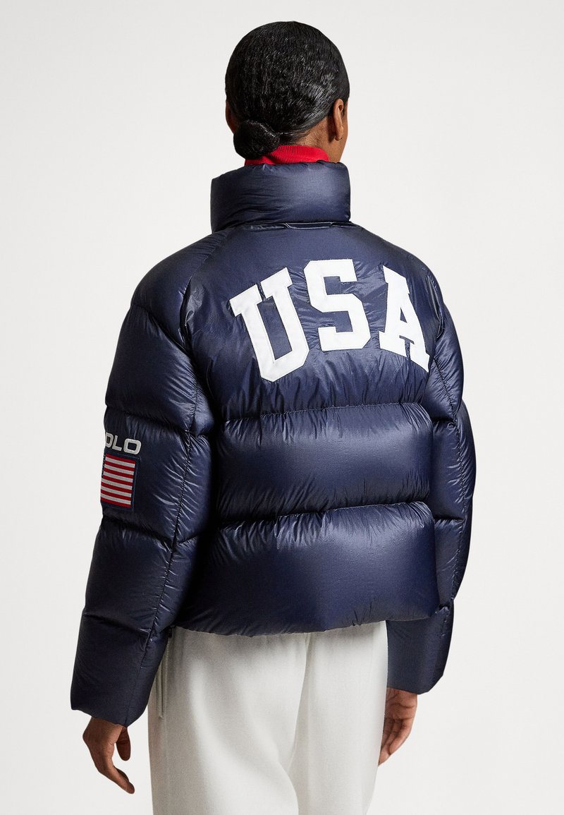 Marinblå vadderad jacka med hög krage, med vita "USA"-bokstäver på baksidan och en lapp med röda ränder på vänster ärm.