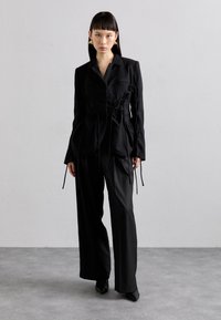 Schwarzer taillierter Blazer mit aufwendigen Schnürdetails an den Seiten, kombiniert mit passenden weit geschnittenen Hosen und spitzen schwarzen Absätzen.