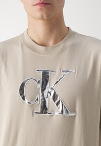 Beige cotton t-shirt featuring a large silver metallic "CK" logo in a glossy finish, with a soft round neck and short sleeves.
