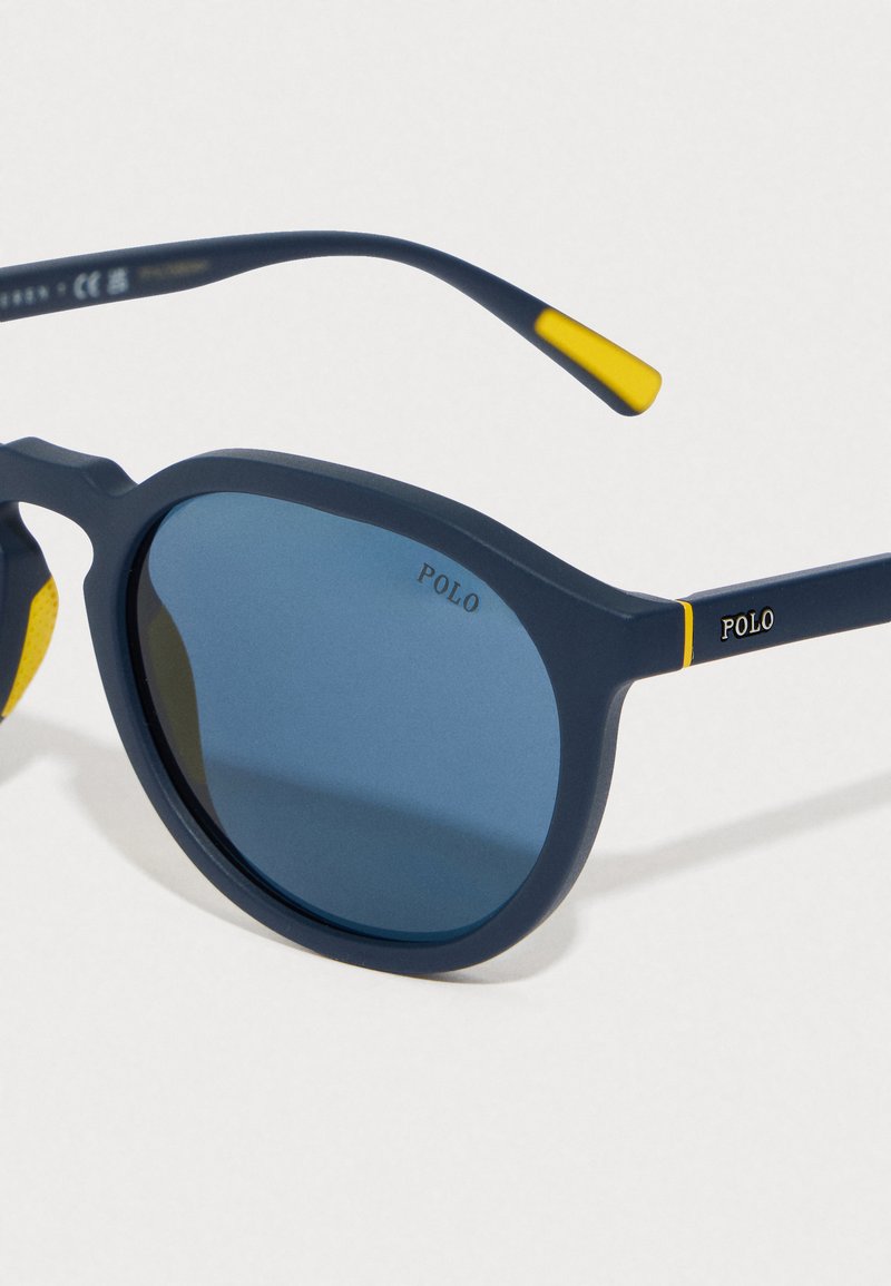 Navy blue sunglasses with round frames, blue tinted lenses, and matte finish. Features yellow accents on the temples and "POLO" branding.
