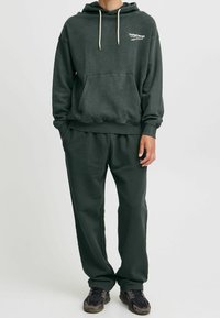 Felpa verde scuro con tasca frontale e cappuccio con cordino, abbinata a pantaloni della tuta coordinati. Il tessuto sembra morbido; le scarpe sono nere con accenti grigi.