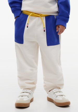 Barn iført hvide joggingbukser med blå lommer og gul snor, kombineret med hvide velcrosneakers med brune såler.