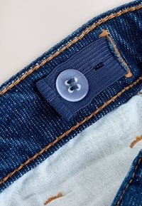 Slet-knap fastgjort til blå elastikbånd på denimstof med orange syning og lys inderforing synlig.