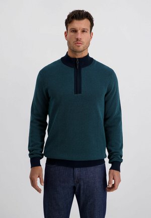 Mann trägt einen türkisfarbenen, texturierten Pullover mit halbhohem Reißverschluss, dunkelblauem Kragen und Bündchen, kombiniert mit dunkelblauen Jeans, steht vor einem einfarbigen Hintergrund.