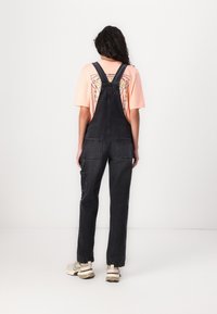 Zwarte denim salopettes met verstelbare banden, twee achterzakken en een zijzak. Draagt over een lichtroze T-shirt met korte mouwen.