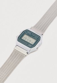 Digitální hodinky se stříbrným obdélníkovým kovovým pouzdrem, zeleným digitálním displejem, mesh páskem a indikátorem voděodolnosti. Zobrazuje čas a datum.