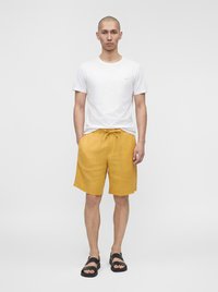 Homme debout les mains dans les poches, portant un t-shirt blanc, un short jaune moutarde et des sandales noires sur un fond gris uni.