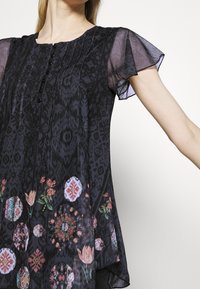 Blusa negra con estampado, con mangas de vuelo transparentes, una fila de botones en la parte delantera y diseños florales y geométricos coloridos cerca del dobladillo.