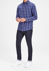 Camisa de cuadros azul y negra, de botones, con mangas largas, combinada con jeans de mezclilla oscuros y zapatillas blancas. Diseño de corte ajustado, estilo casual.