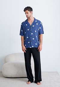 Short-sleeve button-up shirt in blue with white circular pattern and "HUGH" text, paired with loose black pants. Flat surface background.
