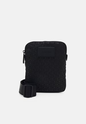 Sac bandoulière compact noir avec motif all-over du logo CK, patch logo à l'avant, fermeture éclair supérieure et sangle ajustable enroulée à côté.