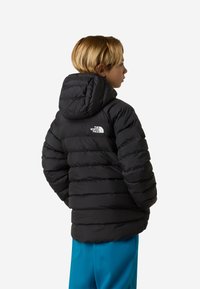 Μαύρο μπουφάν puffer με κουκούλα, διακοσμημένο με οριζόντια ραμμένη σχεδίαση και μικρό λευκό λογότυπο στο επάνω μέρος της πλάτης. Φοράται πάνω από μπλε παντελόνια.