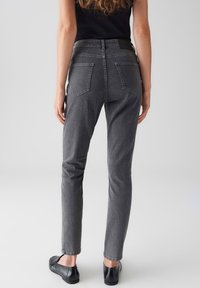 Donna che indossa jeans neri sbiaditi a vita alta e ballerine nere, in piedi con le mani rilassate ai lati su uno sfondo grigio uniforme.