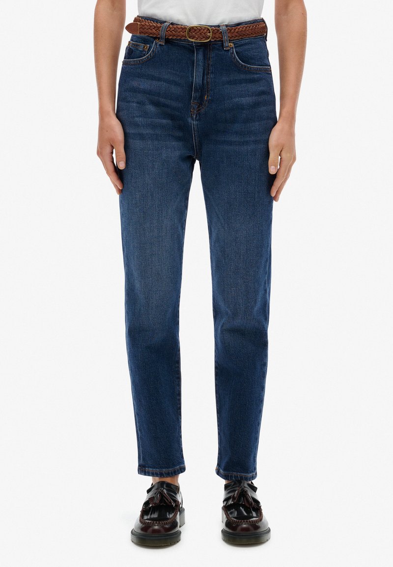 Jeans de mezclilla azul oscuro con cintura alta, pierna recta, cinco bolsillos y un cinturón de cuero trenzado marrón. Combinados con zapatos negros.