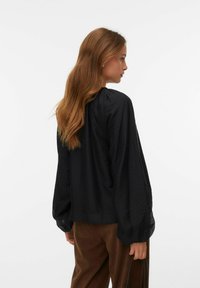 Blusa nera trasparente con maniche lunghe e ampie, scollatura arricciata e texture liscia, abbinata a pantaloni in corduroy marroni.