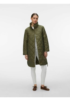 Kobieta w oliwkowozielonym pikowanym płaszczu do kolan, białych spodniach i brązowych sznurowanych butach na jednolitym jasnym tle.