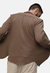 Giacca marrone con una texture, con un leggero allungamento dell'orlo posteriore e il collo, caratterizzata da un tessuto a costine e una vestibilità sartoriale. Dettagli di design minimalisti evidenti.