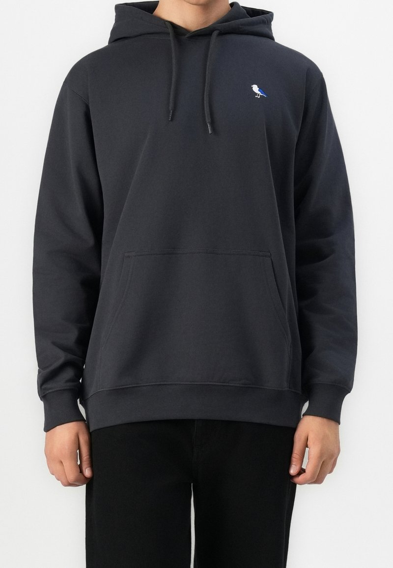 Personne portant un sweat à capuche gris foncé avec un petit logo d'oiseau blanc et bleu sur la poitrine gauche et une poche ventrale.