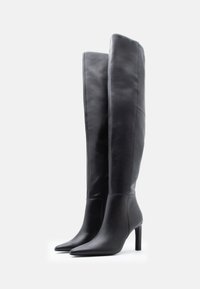 Ζευγάρι ψηλές μαύρες δερμάτινες μπότες με μυτερές μύτες και ψηλά λεπτά τακούνια, τοποθετημένες δίπλα-δίπλα σε λευκό φόντο.