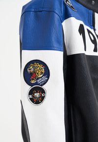 Giacca di pelle con sezioni blu, bianche e nere, dotata di toppe ricamate circolari di una tigre e di un teschio sul braccio sinistro.
