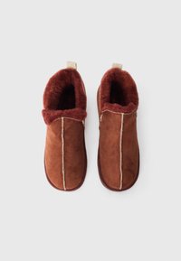 Brown suede slippers with a plush maroon lining, featuring contrasting stitching and a looped pull tab at the back for easy wear.