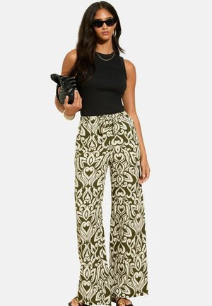 Top senza maniche nero abbinato a pantaloni ampi con un intricato motivo bianco e verde oliva. Gli accessori includono una clutch nera e occhiali da sole scuri.