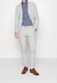Abito grigio chiaro con giacca aderente e pantaloni; abbinato a una camicia blu a fantasia. Scarpe eleganti marroni completano l'outfit.