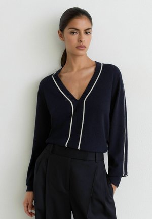 Kvinde med mørkt hår i en lav ponytail iført en sort V-hals sweater med hvide kanter og sorte bukser med høj talje, stående mod en hvid væg.