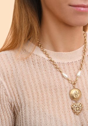 Collier en chaîne dorée avec un pendentif rond et un charme en forme de cœur, rehaussé de perles en goutte blanche, porté sur un pull rose clair.