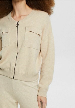 Kvinde iført beige sweatjakke med lynlås og to forlommer samt matchende bukser mod en ensartet lys baggrund.