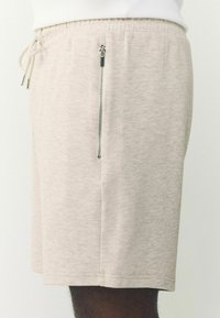 Pantalones cortos beige con cordón y un bolsillo vertical plateado con cremallera visible en el costado, combinados con una parte superior blanca.