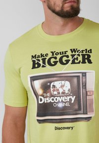 Lichtgroene katoenen T-shirt met een retro tv-graphic waarop de tekst "Make Your World BIGGER" en het logo van "THE Discovery CHANNEL" staan.