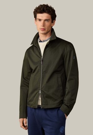 Joven con chaqueta verde oscuro con cremallera sobre suéter beige y pantalones azules, de pie frente a un fondo gris claro liso.