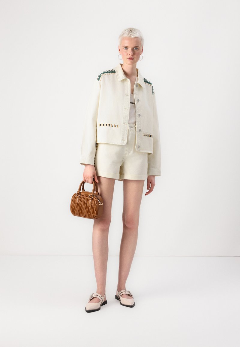 Veste en denim clair avec des accents verts et jaunes, shorts assortis, et un petit sac à main texturé marron ; les chaussures sont roses avec une boucle.