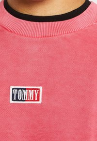 Sudadera rosa con cuello negro acanalado. Presenta un distintivo logo bordado "TOMMY" en rojo y azul sobre un fondo blanco.