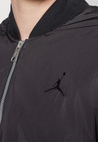Svart nylon bomberjacka med ribbad krage, silverfärgad dragkedja och broderad Jumpman-logotyp på vänster bröst. Lättviktsmaterial.