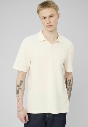 Jeune homme aux cheveux blonds courts portant un polo à manches courtes ivoire et un jean foncé, montrant des tatouages floraux et de papillons sur le bras.